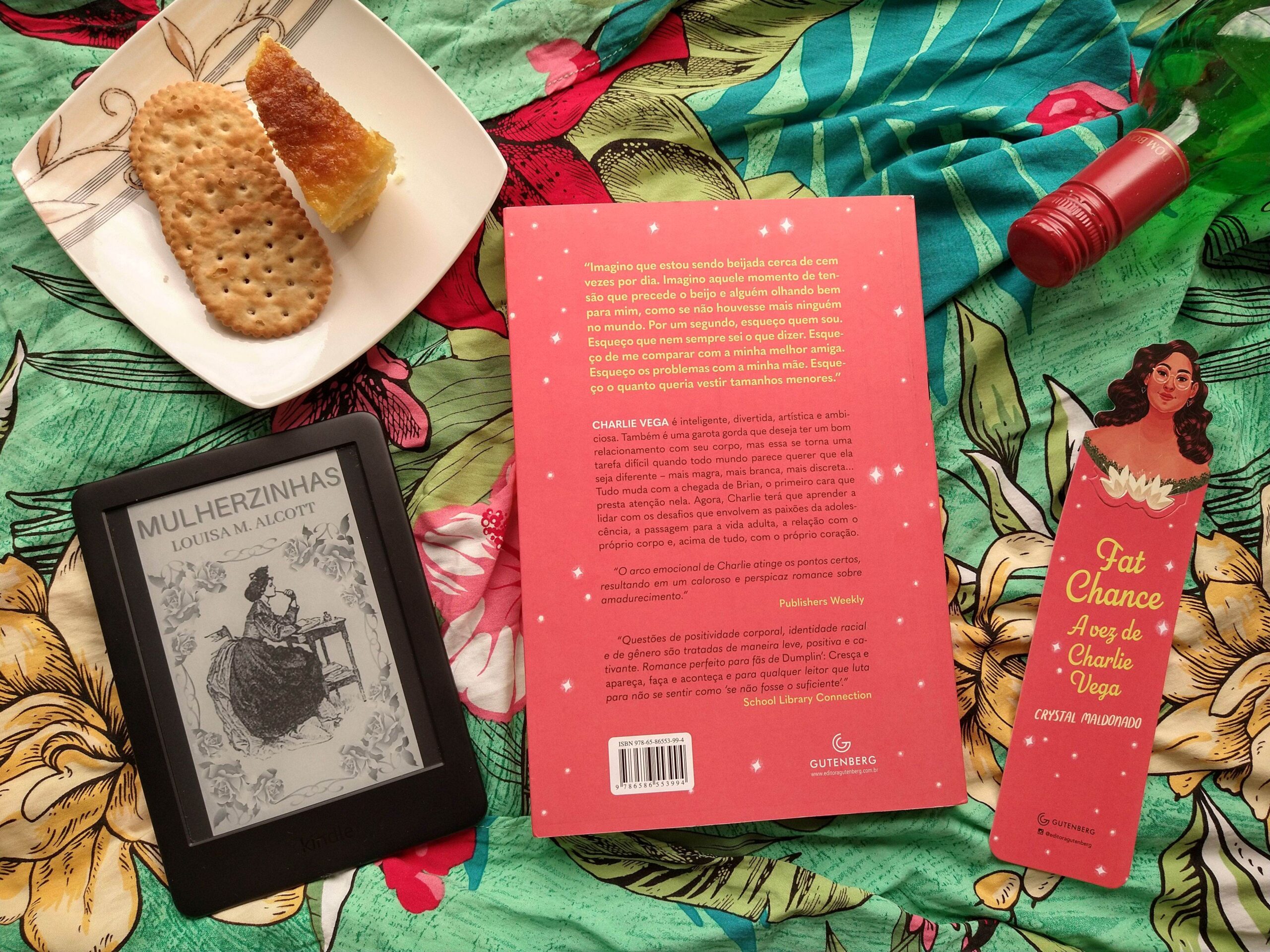 Tecido verde com flores no fundo. Temos o verso do livro com a sinopse, um marcador personalizado da história, uma garra de vinho, um prato com bolo e biscoitos, e um kindle com a capa de "Mulherzinhas".