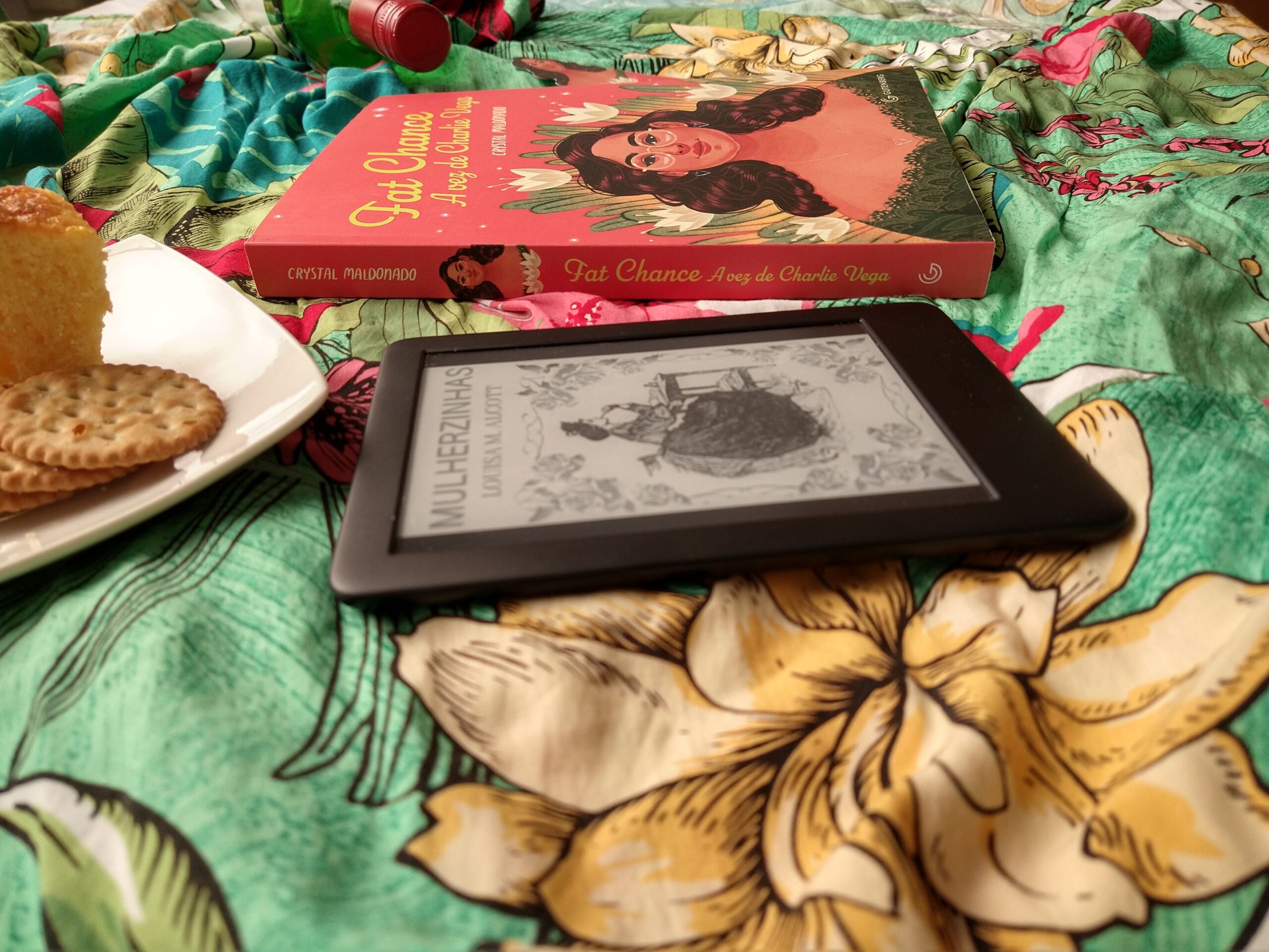 Tecido verde com flores no fundo. Temos a lateral do livro, uma garra de vinho, um prato com bolo e biscoitos, e um kindle com a capa de "Mulherzinhas".