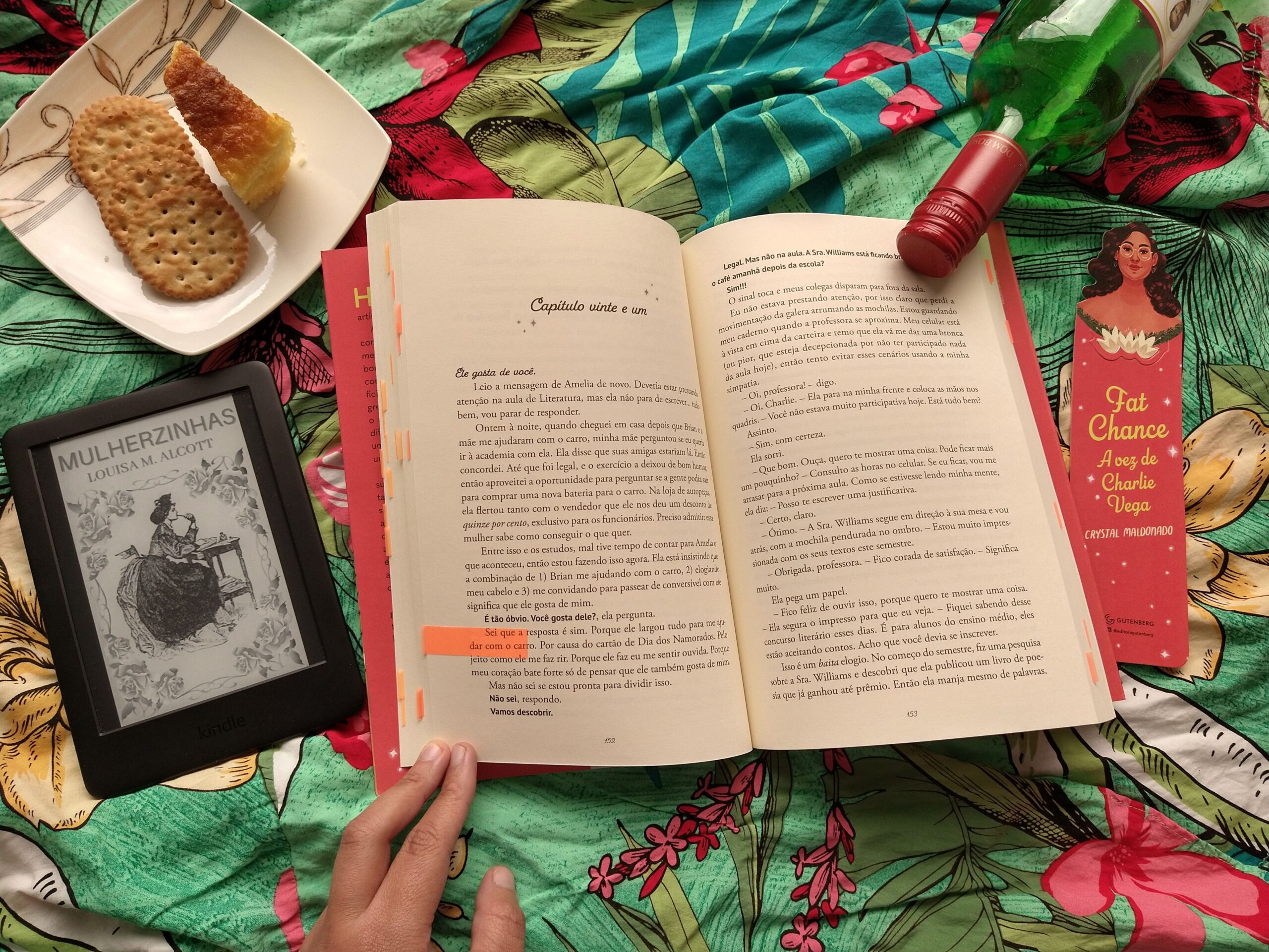 Tecido verde com flores no fundo. Temos o livro aberto no capítulo 21, um marcador personalizado da história, uma garra de vinho, um prato com bolo e biscoitos, e um kindle com a capa de "Mulherzinhas".
